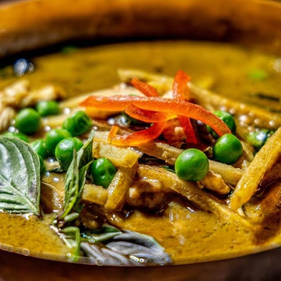 Close-up of creamy Thai green curry with vegetables, green peas, sliced red chilies, and fresh basil in a bowl.