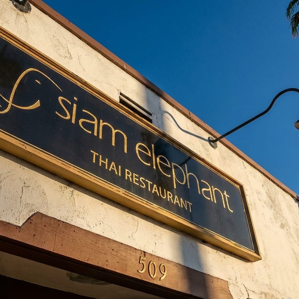 Exterior sign for Siam Elephant Thai Restaurant, featuring a black-and-gold sign, stucco facade, palm trees, and blue sky.