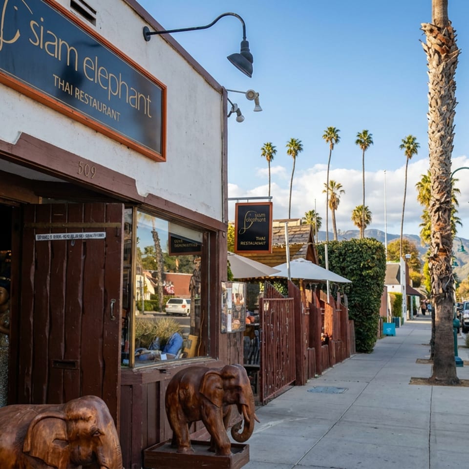 Siam Elephant Thai restaurant storefront with carved elephant statues, outdoor patio, palm-lined street, and mountain backdrop.