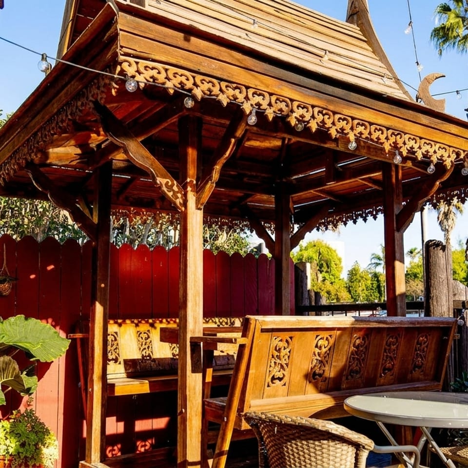 Ornate wooden outdoor dining pavilion with carved benches, patio tables, potted plants, string lights, and palm trees.