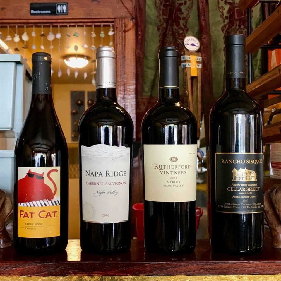 Four wine bottles displayed between carved wooden horse sculptures, with a wine rack, restroom sign, and warmly decorated restaurant interior behind them.
