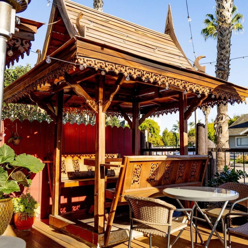Ornate wooden outdoor dining pavilion with carved details, patio tables and chairs, palm trees, and string lights.